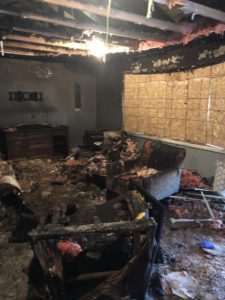 Destroyed living room with soot and smoke damage; All items within, including furniture, is charred.